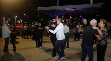 pessoas em duplas estão dançando em pista na orla. Ao fundo, um palco. Não é possível ver os  músicos. #paratodsoverem