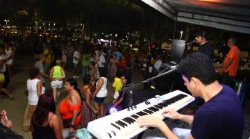 Público dança em frente ao palco do evento ao fundo e, em primeiro plano, músicos tocam em cima do palco. #pracegover