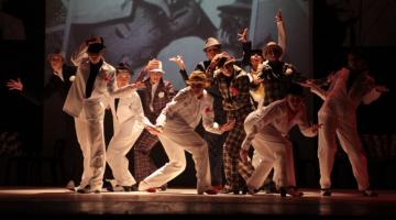 Bailarinos estão no palco fazendo coreografia. Eles usam calças, ternos e chapéus. #Paratodosverem