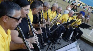 close de músicos sentados lado a lado tocando instrumentos de sopro. eles usam camisetas da mesma cor, em amarelo. Diante deles há estantes com partituras. #paratodosverem