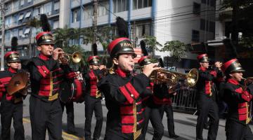 Banda desfila em rua. Músicos uniformizados tocam instrumentos de sopro. #Pracegover