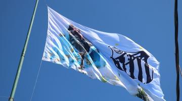 a bandeira no alto do mastro com a arte da santa e o distintivo do clube. #paratodosverem
