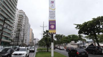 banner afixado em poste em canteiro central de avenida da orla. Carros passam nas duas pistas, indo e vindo. #paratodosverem