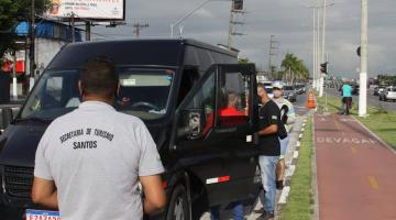 HOMEM COM CAMISETA COM ESTAMPA DO NOME DA SECRETARIA DE TURISMO ESTÁ DE COSTAS, DIANTE DE UMA VAN. A van está de porta abeerta, com duas pessoas ao lado conversando. Eles estão em uma avenida, ao lado de uma ciclovia. #paratodosverem