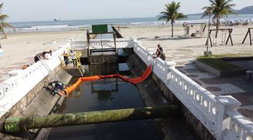 barreira é instalada dentro de canal junto à orla. Dois homens estão de cada lado do canal, sendo um dentro e outro fora. ambos estão segurando as pontas das barreiras. #paratodosverem