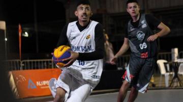 Jogador da equipe de Santos, com uniforme branco, segura a bola de basquete e olha para o alto, com adversário acompanhando de perto. #pracegover