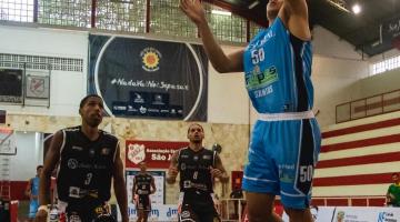 jogador de basquete lança bola ao cesto dentro do garrafão. Ao fundo, dois jogadores do time adversário . #paratodosverem