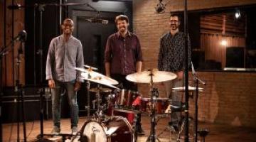 Integrantes do Mário Gaiotto Trio posam para foto atrás de bateria montada em estúdio de música. #paratodosverem