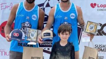 Dois atleta com troféus no pódio e criança na frente #paratodosverem