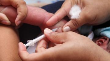 Mãos de adultos seguram criança e aplicam vacina no bracinho. #paratodosverem 