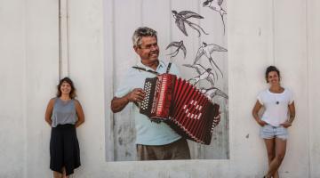 Beatriz e Carolina estão em pé ao lado da imagem de um homem tocando concertina. #Paratodosverem