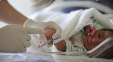 recém-nascido está deitado. Mão com luva segura uma das mãozinhas do bebê. #paratodosverem