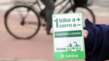 close de mão segurando pequena placa onde se lê uma bike a mais um carro a menos. Ao fundo, uma bicicleta passando. #paratodosverem