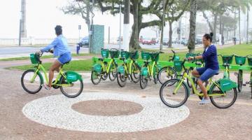 duas mulheres estão montadas em bicicletas do bike santos ao lado de estação.  A estação fica em uma faixa de jardim da orla. #paratodosverem