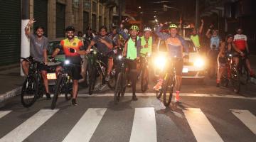 ciclistas param diante de faixa de pedestre para foto. É noite. Eles acenam. #paratodosverem