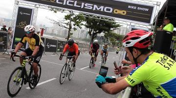 Ciclistas pedalam na avenida enquanto ciclista tira foto pelo celular do lado de fora da prova. #pratodosverem