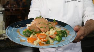 mãos do chef junior monteiro seguram prato com peixe frito, legumes e arroz. #paratodosverem