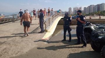 três guardas municipais e quadriciclos estão ao lado de ponte que atravessa canal na faixa de areia. Pessoas cruzam a ponte. #paratodosverem