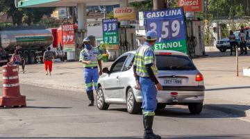 Veículo para próximo à guia de calçada diante de agente da CET. Outro agente está em primeiro plano olhando para o carro. Ao lado direito há um posto de combustível.. #Paratodosverem