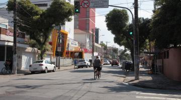 trecho de rua com calçadas, carros estacionados dos dois lados e uma mulher de bicicleta indo adiante da foto. #paratodosverem