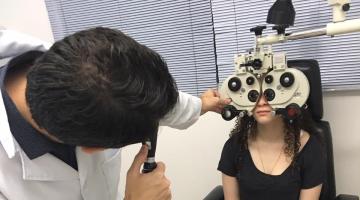Oftalmologista em pé examina aluna sentada com aparelhos nos olhos. #pracegover