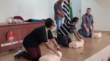 três pessoas estão ajoelhadas no chão massageando peito de boneco que simula uma pessoa. Uma dessas pessoas é o bombeiro que orienta o procedimento. #paratodosverem 