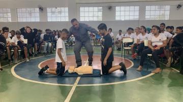 bombeiro está no centro de uma quadra, entre dois meninos que olham para um boneco deitado no chão. #paratodosverem