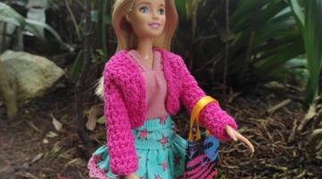boneca barbie vestida com roupa casaco cor de rosa e saia azul e rosa. Ela tem uma bolsa pendurada no braço esquerdo. #paratodosverem
