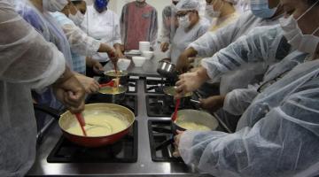 várias pessoas estão mexendo em panelas, fazendo receita de brigadeiro, em torno de um fogão industrial. #paratodosverem