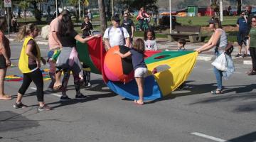 crianças e adultos estão na avenida da orla brincando com grandes recortes de tecidos e bolas coloridas. #paratodosverem