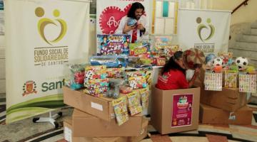 vários brinquedos dentro de caixas e sobre mesas. Um homem está atrás da mesa e à frente de banners. Ele ri. Á frente, uma mulher está dentro de uma caixa e cobre parte do rosto com um chapéu.#paratodosverem