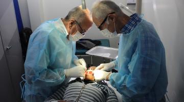 dois cirurgiões estão em pé atendendo um homem que está deitado de boca aberta. #paratodosverem
