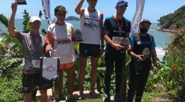 Atletas no pódio erguem troféu com paisagem de fundo #paratodosverem