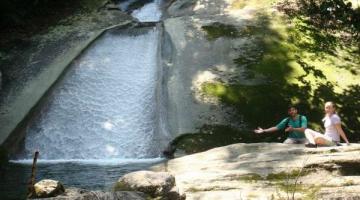 cachoeira com piscina natural na parte inferior. Do lado direito, duas pessoas estão sentadas sobre pedras. Há vegetação em torno. #paratodosverem