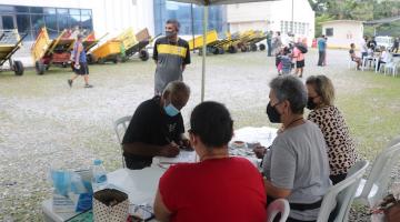 homem está sentado diante de mesa, sendo atendido por três pessoas. Ao fundo, vários carrinhos de coletores. #paratodosverem