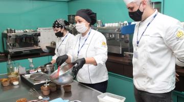 duas mulheres e um homem vestidos de culinaristas estão atrás de mesa. As duas estão preparando um prato. #paratodosverem