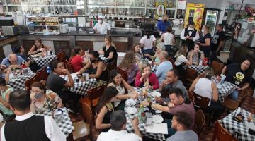 Restaurante lotado de clientes sentados em torno de mesas com toalhas estampadas. #paratodosverem