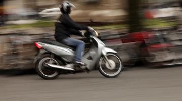 Imagem de homem em moto prateada passando por via pública com imagem de fundo desfocada. #pratodosverem