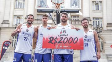 Atletas mostram cheque de R$ 2 mil da premiação em frente à cesta de basquete, com Prefeitura ao fundo. #pracegover