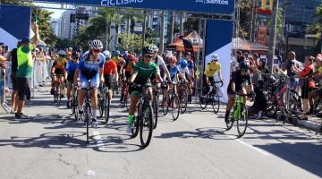 ciclistas estão iniciando a prova. Há um portal ao fundo onde se lê no alto Santista de ciclismo. #paratodosverem 