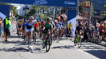 ciclistas estão fazendo largada de prova. Pórtico ao fundo tem escrito Campeonato Santista de Ciclismo. #paratodosverem