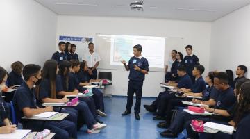 aprendizes estão sentados em sala. Alguns estão em pé. Um deles, o principal, no meio da sala, fala ao grupo. #paratodosverem