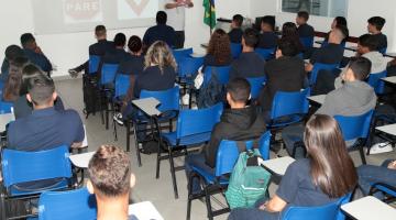 sala está cheia de jovens sentados assistindo palestra. Homem fala ao fundo. No telão que está atrás dele se vê dois sinais de trânsito. #paratodosverem