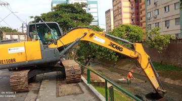 escavadeira está na ocupando parte da rua e calçada e com o braço pra dentro do canal removendo detritos. Ao fundo, no canal, há um homem também coletando detritos. #paratodosverem