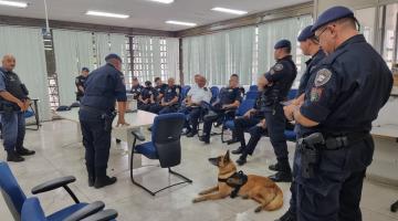 Guarda municipais estão em sala. Alguns estão em pé e outros sentados. Pastor alemão está no chão. #paratodosverem