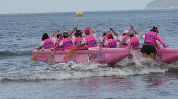 Canoa havaiana na cor rosa ocupada com mulheres vestidas de rosa ingressa no mar. #pracegover