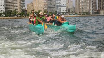 mulheres remam na água dentro da canoa. Há movimentação de ondas. #paratodosverem