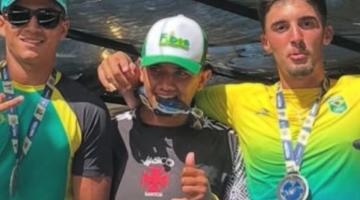 trés atletas posam para foto abraçados. O do meio segura uma medalha