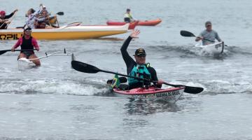 canoísta está dentro do caiaque na beira do mar. Com uma mão ele segura o remo e outra está erguida para o alto. #paratodosverem