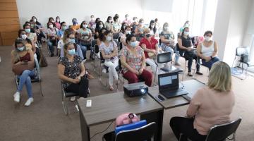 profissionais de saúde estão sentados em sala, em cadeiras distantes umas das outras. Ele olham para mulher que está sentada diante uma mesa com um notebook. #paratodosverem